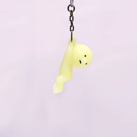 The adorable Smiski Keychain - Grabbing Key Figure, with its charming yellow hue, glows in the dark as it hangs from a chain against a simple backdrop.