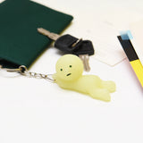 A small, yellow Smiski Grabbing Key Figure keychain rests beside a green wallet and car keys on a white surface. This cute accessory from Smiski, with its glow-in-the-dark feature, adds a whimsical touch to your everyday essentials.