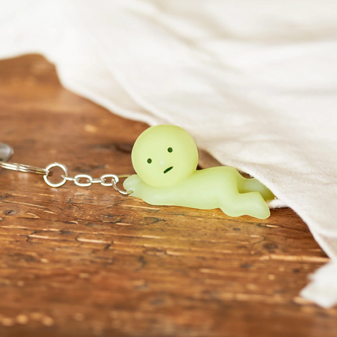 The Smiski Keychain - Grabbing Key Figure, by Smiski, is a charming accessory featuring a small, green, human-like figurine lying on a wooden surface partially covered with white fabric. It even glows in the dark for an extra whimsical touch.