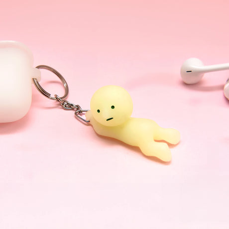 A small, pale yellow Smiski Keychain - Grabbing Key Figure from the Smiski brand rests on a pink surface next to a white earbud and a white container. This adorable, glow-in-the-dark charm brings whimsy and charm to any collection.