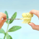 A pair of hands holds a distinctive Smiski Keychain - Pulling Key Figure, featuring three linked small yellow figures. Renowned for their glow-in-the-dark quality by the Smiski brand, this keychain ensures you won't misplace your keys again. The soft blur of a green plant serves as an ideal backdrop.