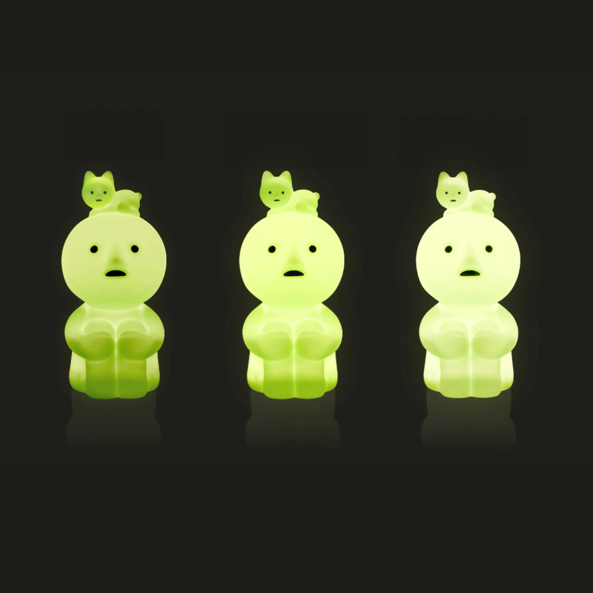 Against a dark backdrop, three small, glowing green figures with wide eyes and round faces stand, each adorned with a tiny creature on its head. These Smiski Touch Light companions by the brand Smiski make the perfect touch-activated nightlights for soothing evenings.