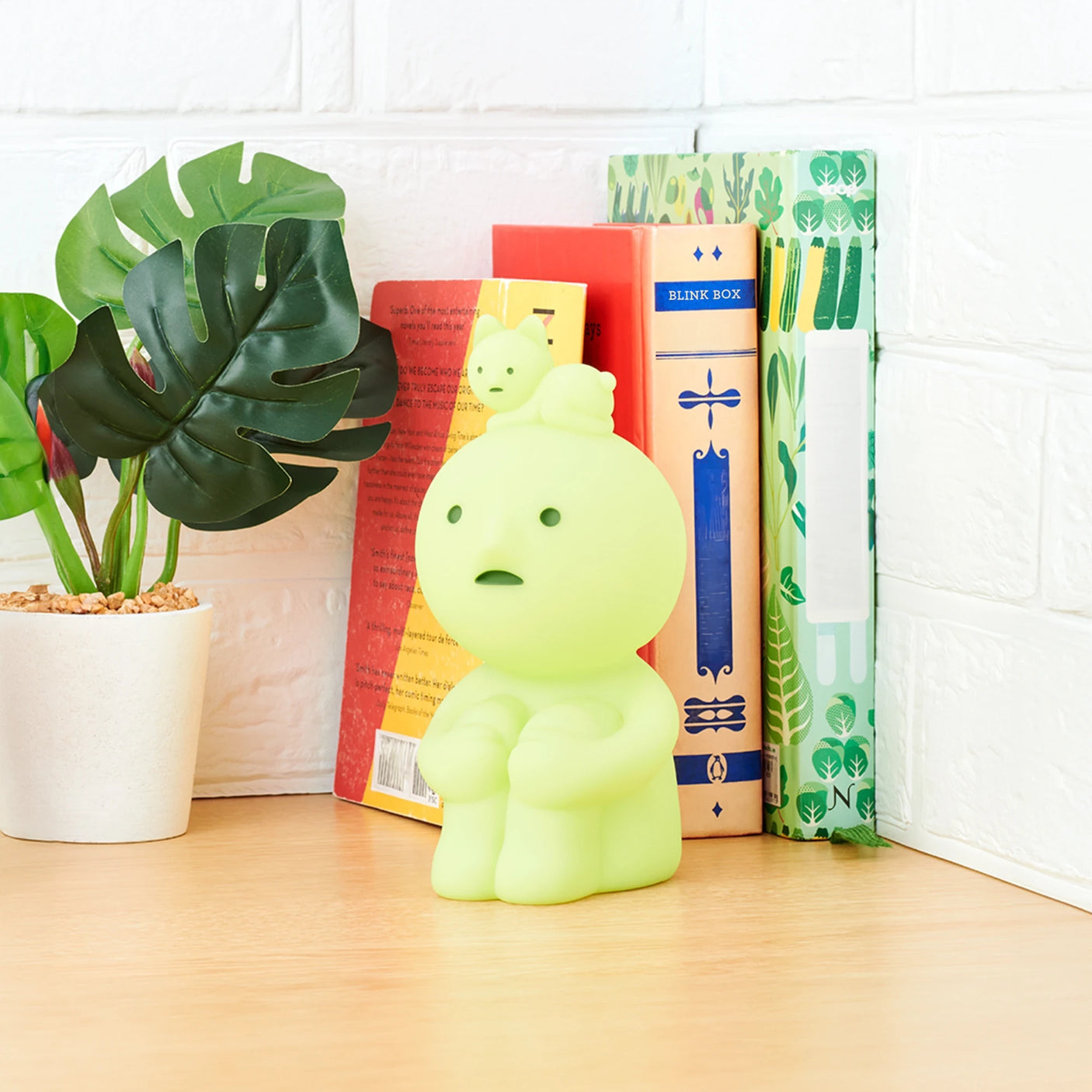 A Smiski Touch Light, featuring a green glow and a small figure atop it, rests on a wooden surface next to books and a potted plant. This whimsical, touch-activated nightlight adds charm to your space.