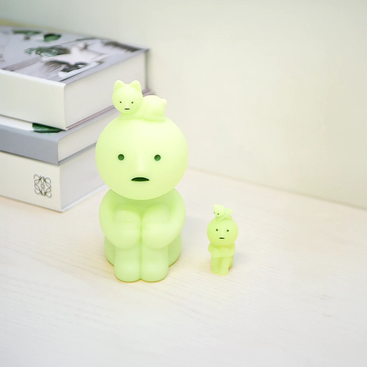 Two Smiski Touch Lights, featuring green figurines with small animals on their heads, sit beside stacked books on a desk, casting a soft glow reminiscent of a whimsical nightlight.