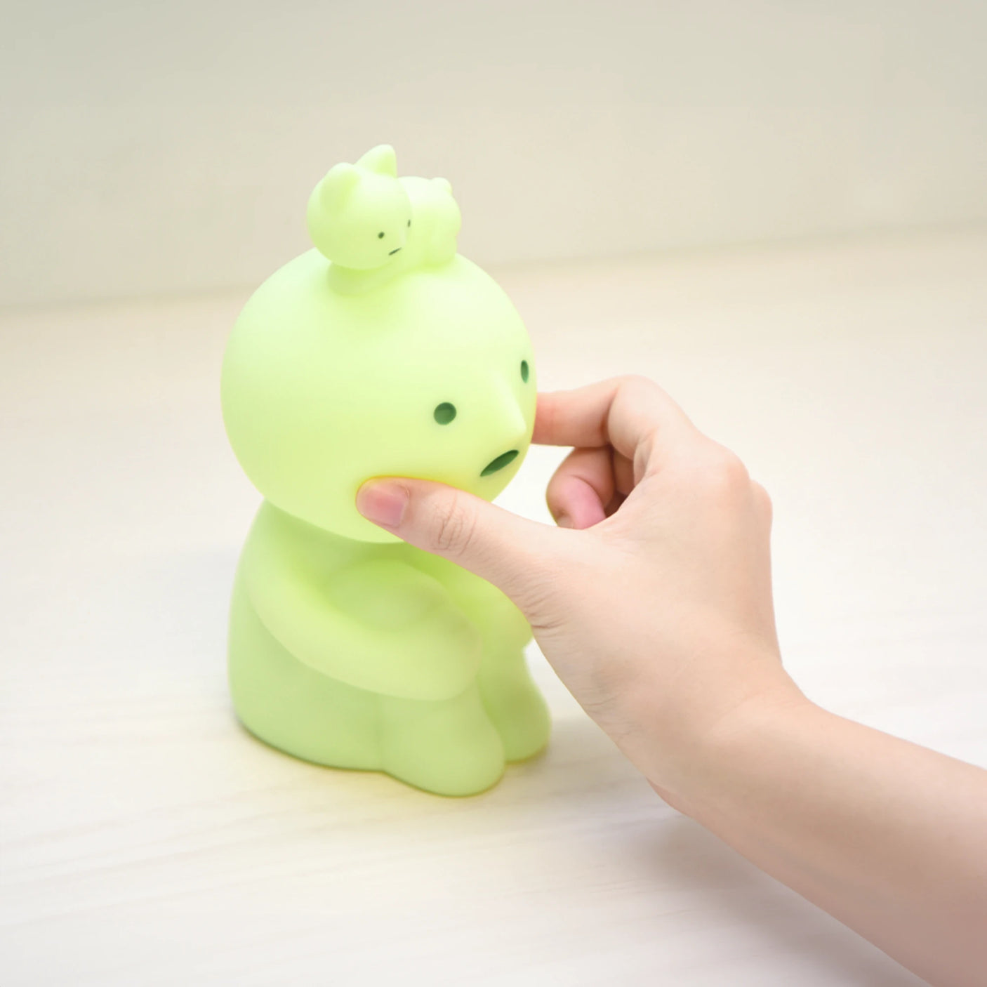 A hand squishes the cheek of a small, green, round-headed figurine that resembles a Smiski Touch Light nightlight by Smiski, with a smaller figure perched on its head, placed on a light surface.