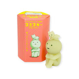 A glow-in-the-dark Smiski figure from the Smiski Living Series stands with arms crossed, with two smaller figures perched on its head. It is positioned next to its box, which features Japanese text and branding, as part of the entertaining Smiski Living Series - Blind Box collection.