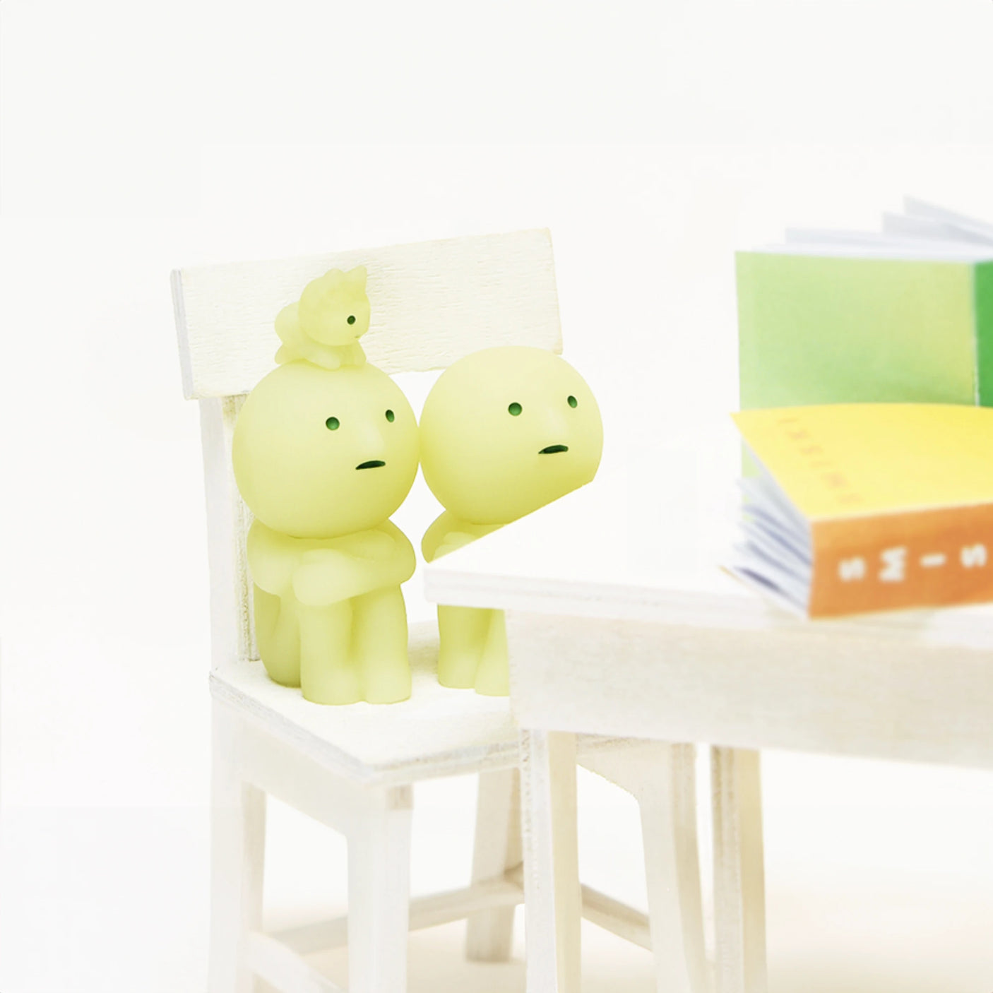 Two small, round, pale green Smiski figures from the Smiski Living Series - Blind Box are seated on a white chair, with a tiny bird perched on one figure's head. They gaze at open books on a white table, exuding a whimsical aura. It's like uncovering the mystery of a lively Blind Box in your own space.