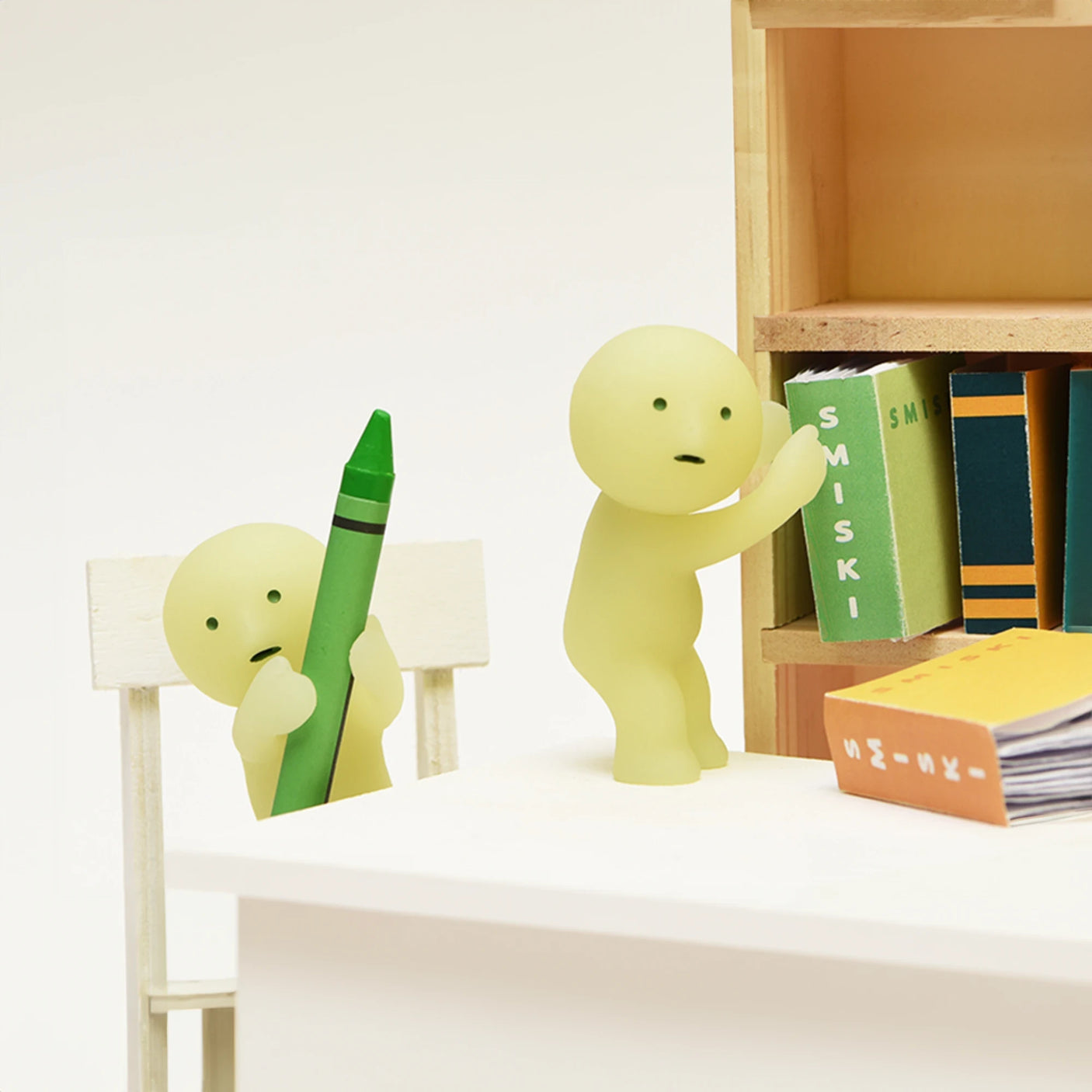 In the Smiski brand's Living Series - Blind Box, two small green glow-in-the-dark figures are featured, one holding a crayon while the other reaches for a book on a shelf.