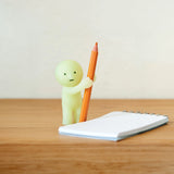 A small figure from the Smiski Living Series by Smiski, featuring a green character holding an oversized pencil, stands next to a notepad on a wooden surface. This glow-in-the-dark figurine adds a whimsical and mysterious touch.