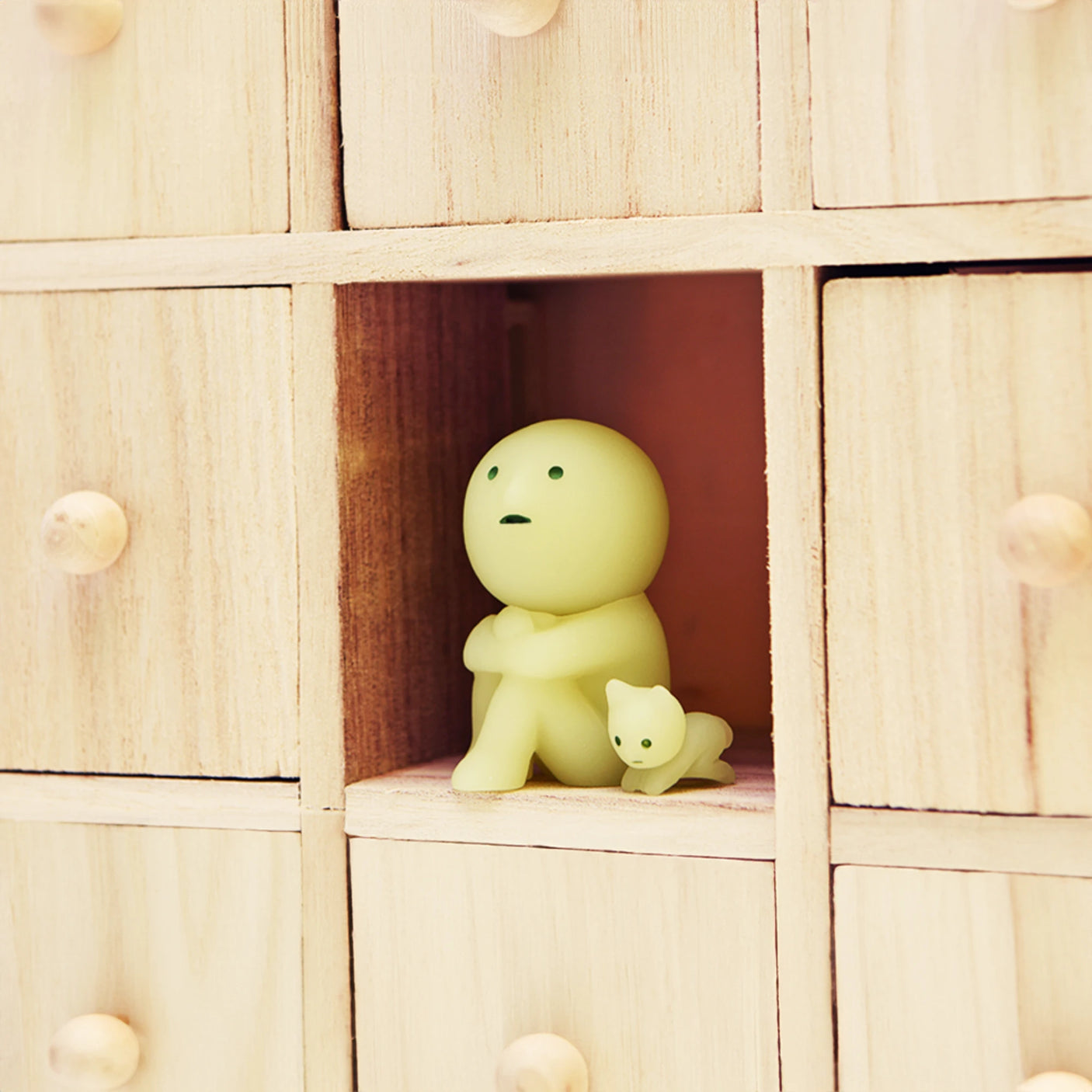 A tiny, rounded figure from the Smiski Living Series by Smiski and a cat figurine rest together in an open wooden cubby, surrounded by closed drawers. It's as if they're poised to embark on an adventure once they break free from their Smiski Living Series - Blind Box mystery.