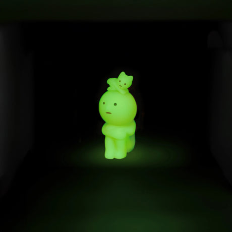 A glowing green figure from the Smiski Living Series by Smiski, featuring a cartoon design and a small cat on its head, stands in the dark.