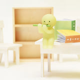 A small, green, spherical figure from the Smiski Living Series - Blind Box by Smiski is positioned on a white table, playing a green flute. Open books are scattered nearby while an empty bookcase looms in the background, enhancing the mystery of this Glow-in-the-Dark scene.