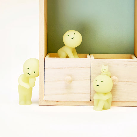 Figurines from the Smiski Living Series - Blind Box, featuring a small, glow-in-the-dark design, peek out from a wooden box with drawers, creating a delightful mystery against a light background.