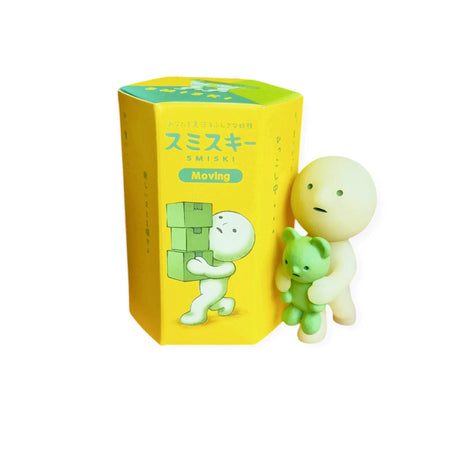 A glow-in-the-dark toy figure holding a green teddy bear stands next to a yellow and green box labeled "Smiski Moving Series - Blind Box" with Japanese text, adding charm to any collection of Smiski figurines.