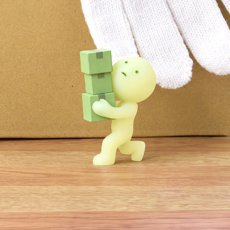 A figurine from the Smiski Moving Series - Blind Box, featuring a small green character balancing three stacked cubes as a large white-gloved hand hovers above, is ready to glow in the dark. The collectible toy by Smiski is set against a wooden surface and a beige background.