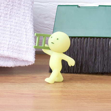 A Smiski Moving Series Blind Box collectible toy from the Smiski brand, featuring a mysterious figure with a green ladder, stands on a wooden surface amidst white textured fabric and the bristles of a push broom. This whimsical character captivates onlookers as it gets ready for its next glow-in-the-dark adventure.