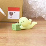 A small, glow-in-the-dark figurine from the Smiski Moving Series - Blind Box by Smiski holds a green box labeled "fragile" on a wooden surface, with a cardboard box and bubble wrap in the background.