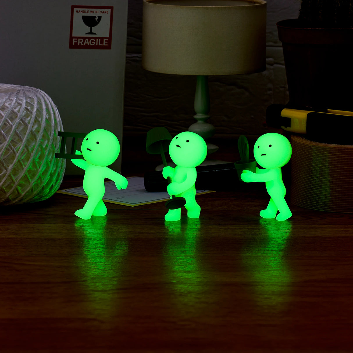 Three Smiski Moving Series figures from a Blind Box clutch gardening tools as they line up on a wooden surface, nestled among a rope and various office supplies.