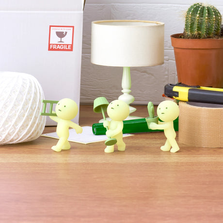 A Smiski Moving Series - Blind Box reveals three small yellow figurines, each with a unique accessory: one holds a ladder, another a shovel, and the third a potted plant. These glow-in-the-dark collectibles from Smiski rest on a wooden surface close to a lamp, a cactus, and a box marked as fragile.