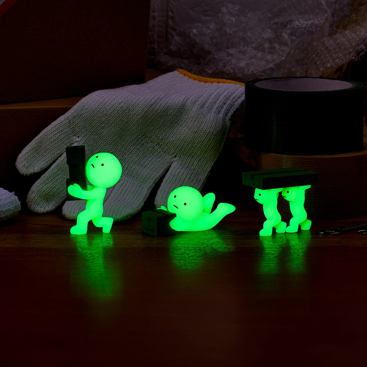 Four Smiski Moving Series glow-in-the-dark stick figure toys are striking various poses on a table, accompanied by a pair of white gloves and rolls of tape.