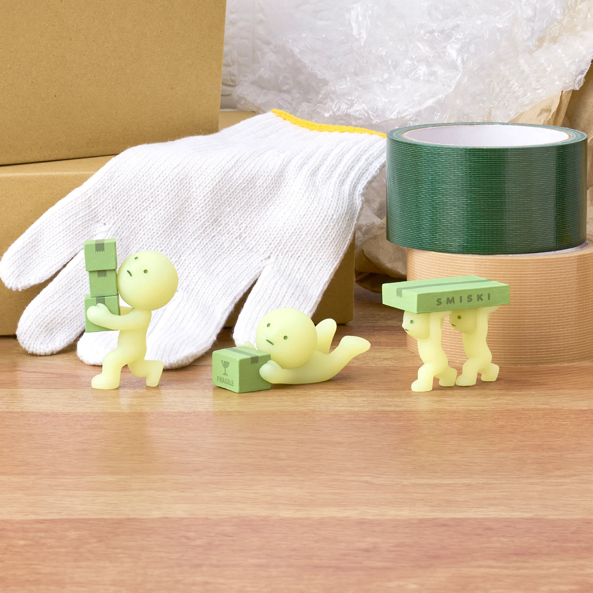 Three figurines from the Smiski Moving Series - Blind Box by Smiski are engaged in playful activities on a wooden surface, interacting with mini green boxes. These collectible toys feature glow-in-the-dark capabilities. Nearby, packing supplies like gloves and rolls of tape add to the scene.