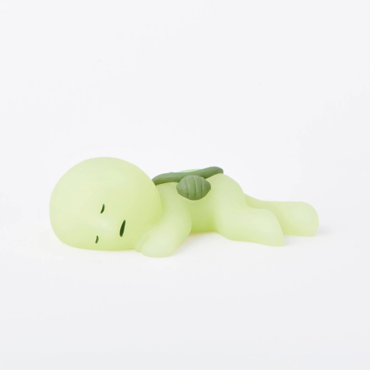 A small, green, cartoonish figure with closed eyes is lying down with a backpack on its back, part of the Smiski Museum Series - Blind Box by Smiski. These glow-in-the-dark figures are perfect for adding a touch of creativity to your space.