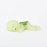 A small, green, cartoonish figure with closed eyes is lying down with a backpack on its back, part of the Smiski Museum Series - Blind Box by Smiski. These glow-in-the-dark figures are perfect for adding a touch of creativity to your space.