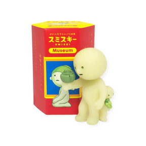 Two small, glow-in-the-dark figures stand near a vibrant red and yellow "Smiski Museum Series - Blind Box." One creative companion holds a small pot, while the other balances a green object on its back, making them intriguing additions to this Smiski collection by the brand.