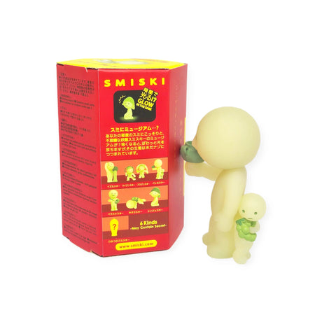 A small glow-in-the-dark figurine from the Smiski Museum Series - Blind Box by Smiski, featuring a baby on its back, stands next to a vibrant red and yellow box decorated with illustrations of these imaginative companions.