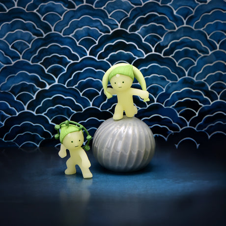 Two figures with leaf-like hair, which glow in the dark, are positioned on and beside a textured round object. They are set against a patterned blue backdrop, reflecting the style of the Smiski Museum Series - Blind Box by Smiski.