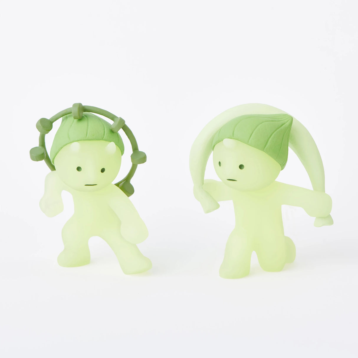 Two small Smiski figurines from the Museum Series, featuring round bodies and leaf-topped heads. The left figurine has a hoop around its head, while the right one sports a single leaf like a hat. These creative companions glow-in-the-dark!