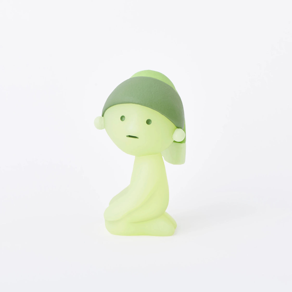 A small, green figurine with a rounded body and a hat sits on a flat surface against a plain white background, featured within the Smiski Museum Series - Blind Box by Smiski.