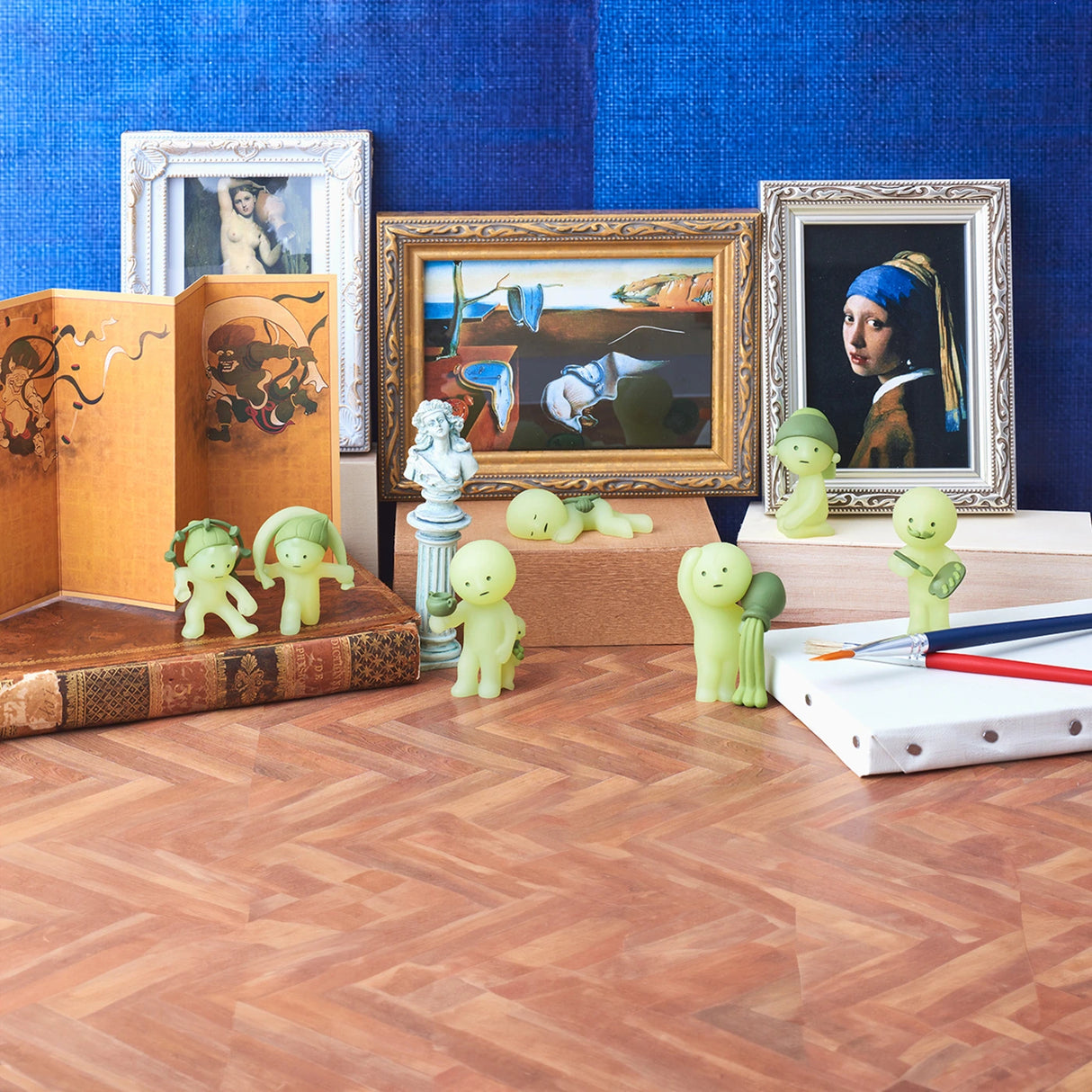 The Smiski Museum Series - Blind Box figures from Smiski are artfully arranged around books, pencils, and prints on a wooden surface with a blue backdrop, serving as creative companions in this imaginative setting.