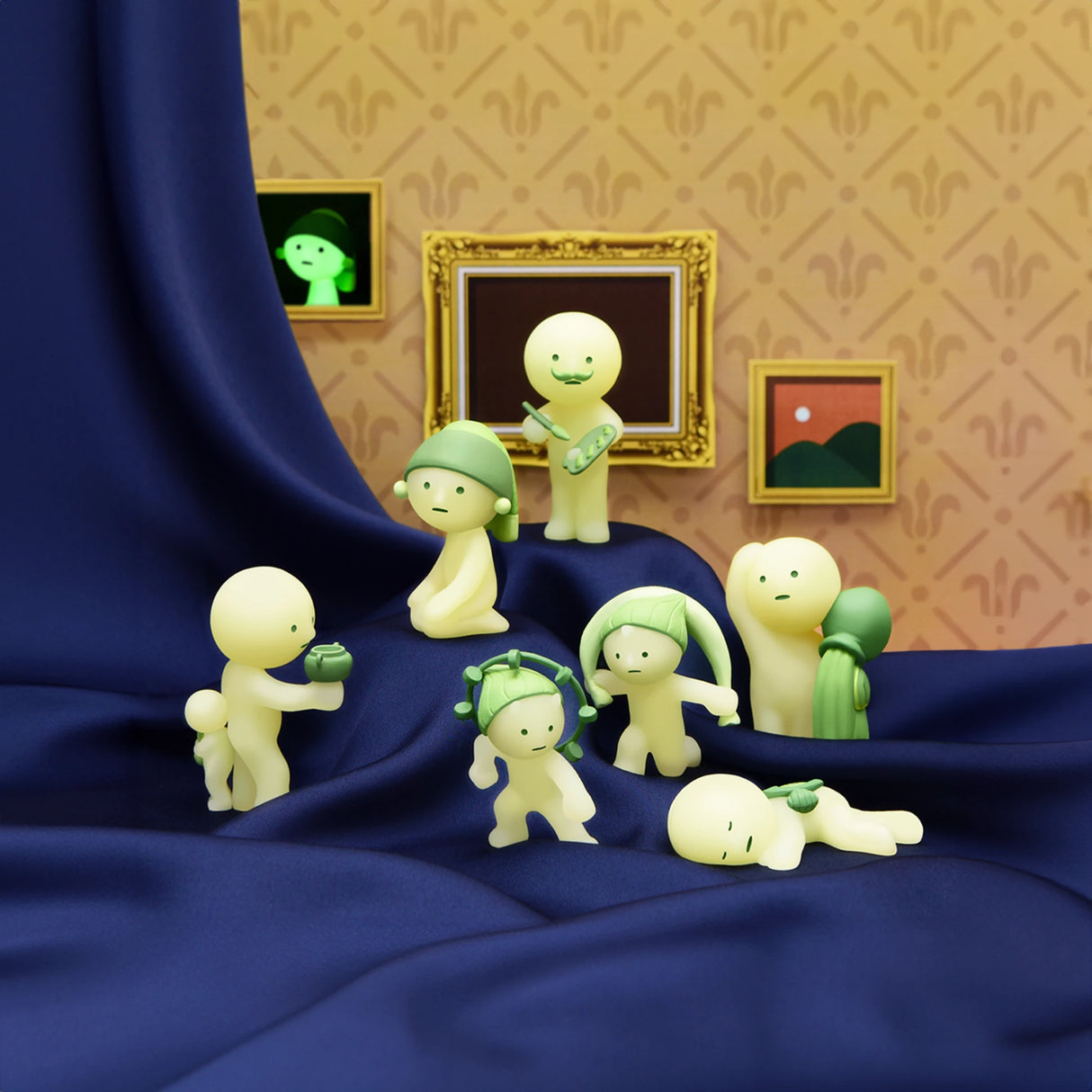 A selection of small, green-tinted figurines adorned with leaf and plant elements from the Smiski Museum Series - Blind Box by Smiski are arranged on a blue cloth, with framed art hanging in the background. These glow-in-the-dark figures make for imaginative companions in any creative environment.