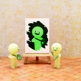 Two small, glow-in-the-dark figures hold paintbrushes as they stand by an easel displaying a painting of a green figure with a smiling face on a textured background. These artistic companions are from the Smiski Museum Series - Blind Box by Smiski.