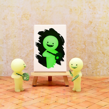 Two small, glow-in-the-dark figures hold paintbrushes as they stand by an easel displaying a painting of a green figure with a smiling face on a textured background. These artistic companions are from the Smiski Museum Series - Blind Box by Smiski.