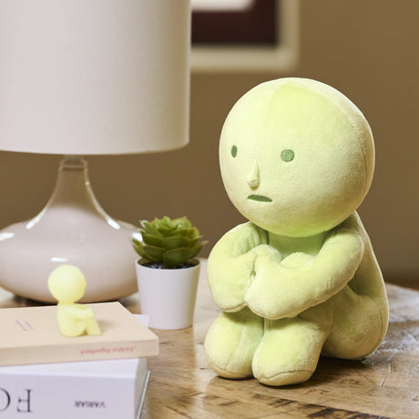 Soft light green Smiski Plush figure sitting with arms hugged around knees on wooden table near lamp and small succulent plant