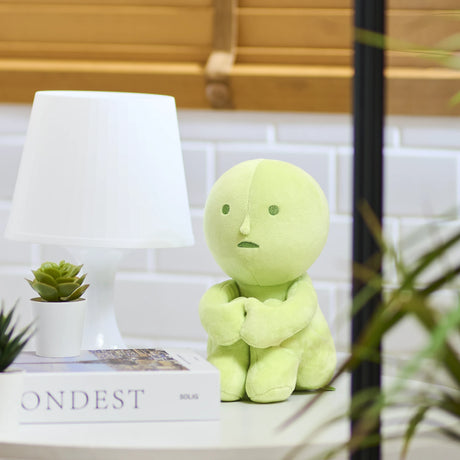 Light green Smiski Plush figure sitting with arms wrapped around knees on white countertop near lamp and plant