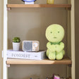 Lime-green Smiski hugging knees plush figure standing on shelf, stitched sleepy face