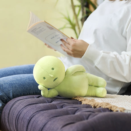 Soft green Smiski Plush figure reclining on side with neutral expression on dark cushioned surface next to person reading book
