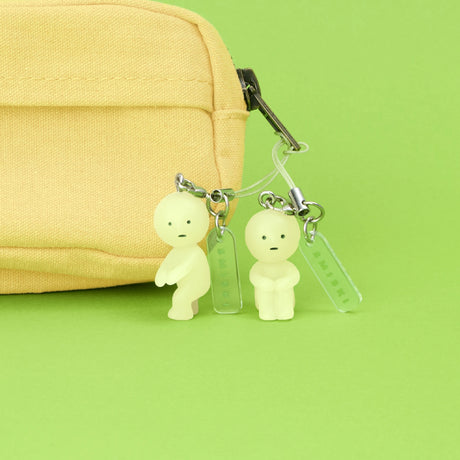 Two miniature glow-in-the-dark Smiski figures keychains attached to zipper of yellow fabric pouch on green background