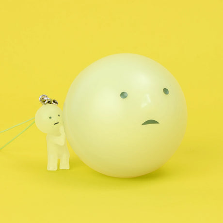 Miniature glow-in-the-dark Smiski figure keychain with worried expression standing next to large glowing orb on yellow background