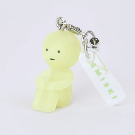 Miniature glow-in-the-dark Smiski figure keychain with tag attached to metal clasp on plain white background