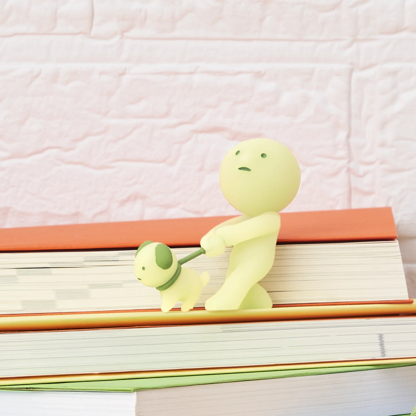 The Smiski Sunday Series - Blind Box by Smiski features a small, pale yellow figurine of a person walking a dog on books with a brick-patterned background—ideal for collectors and glow-in-the-dark enthusiasts.