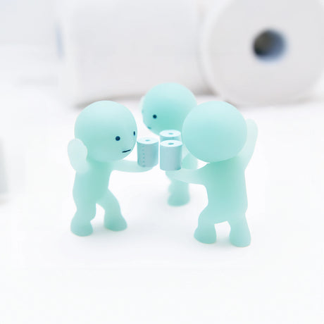 A trio of diminutive, light green glow-in-the-dark figures from the Smiski Toilet Series - Blind Box by Smiski hold tiny toilet paper rolls, positioned on a white surface with larger rolls in the background. Ideal for fans of whimsical mini collectible figures.