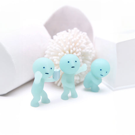 Three small blue Smiski figurines from the Toilet Series stand on a white surface, each featuring simple facial expressions. One figure is holding an item that looks like a cup. A textured white background adds to the scene's appeal, making these Smiski collectibles truly captivating with their whimsical charm.