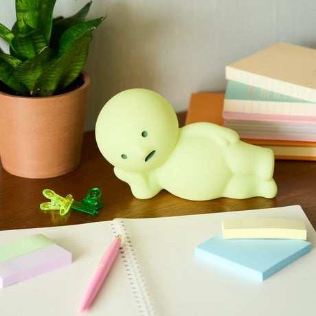 Light green reclining character figurine leaning on one arm on wooden desk with stationery and books nearby