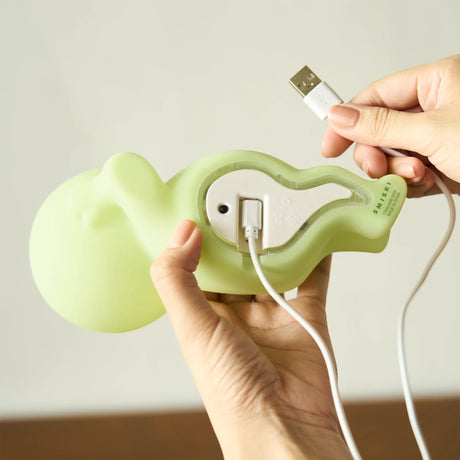 Hands holding light green character-shaped device with USB cable being plugged into its back panel