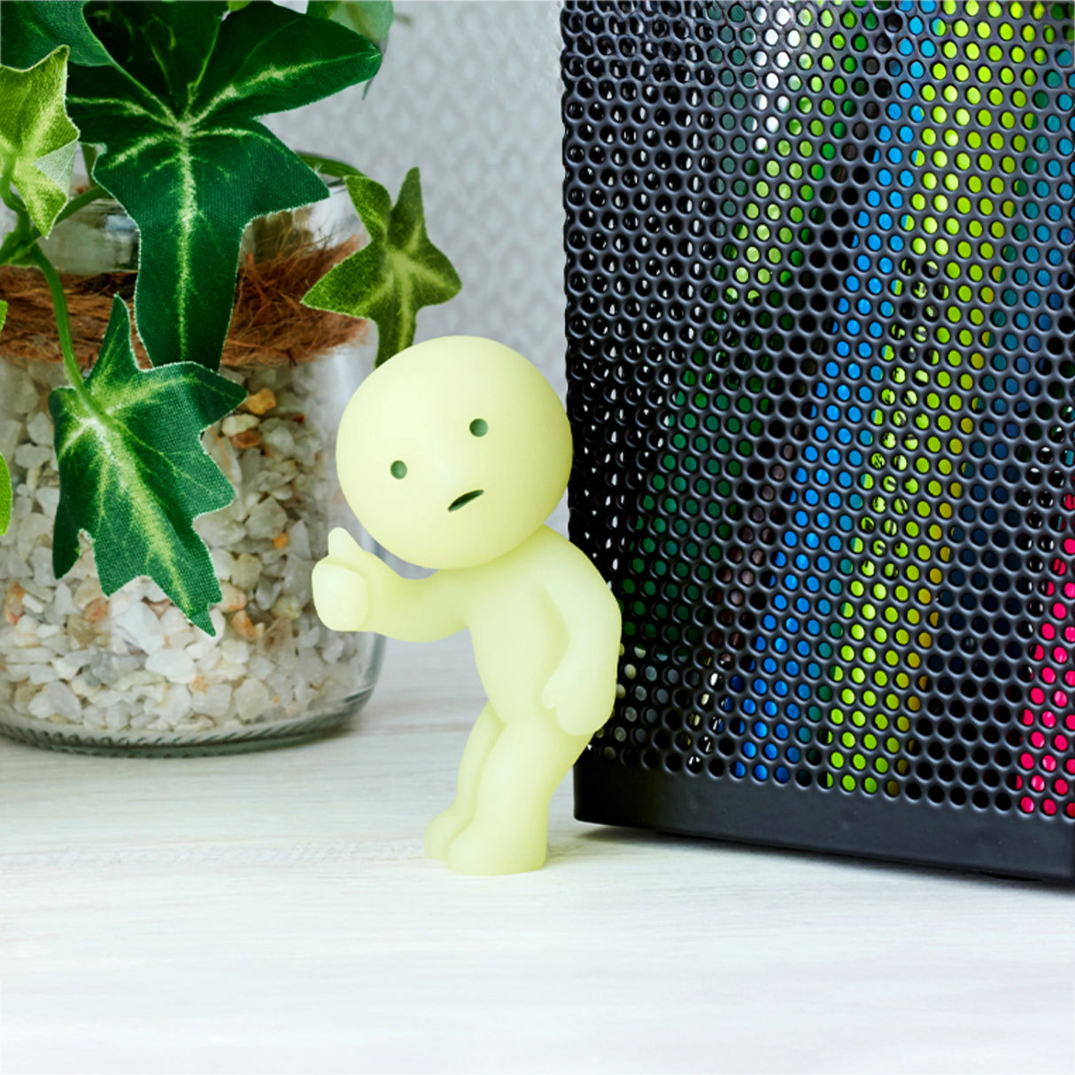 A small, yellow figure from the Smiski At Work Series by Smiski leans against a textured black surface next to a plant with green leaves in a pot filled with pebbles. The enchanting charm of this glow-in-the-dark collectible is concealed within each blind box.