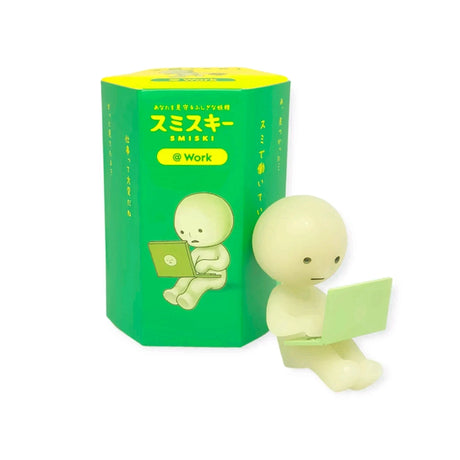 A photo of a Smiski figure from the Work Series, holding a laptop in its lap and depicted as working, placed next to its Blind Box packaging.